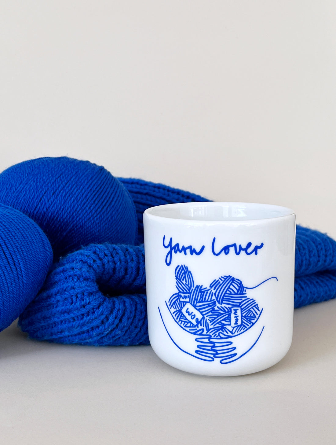 Becher ohne Henkel. Weiß mit Schriftzug Yarn Lover und Motiv Hände umarmen mehrere Wollknäuel in blau.
