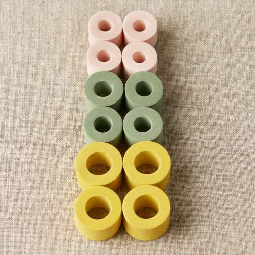 Je 4 Maschenstopper in Gelb, Grün und Rosa auf neutralem Hintergrund