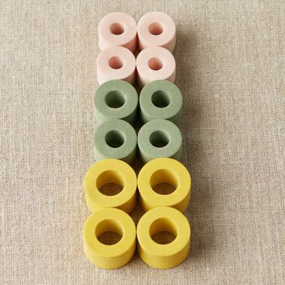 Je 4 Maschenstopper in Gelb, Grün und Rosa auf neutralem Hintergrund